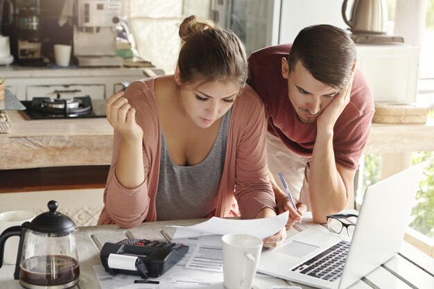 Young married couple with many debts doing paperwork together, reviewing their bills, planning family budget and calculating finances at kitchen table with papers