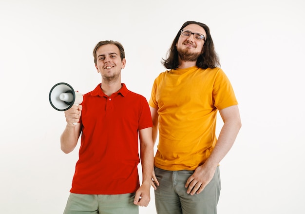 Young men weared in LGBT flag colors on white wall. Caucasian male models in bright shirts. Look happy, smiling and hugging. LGBT pride, human rights and choice concept. Holding mouthpiece.