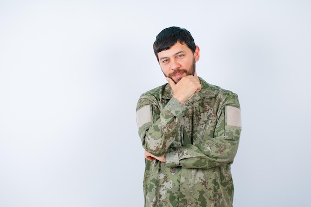 Free photo young military man is looking at camera by holding hand on chin on white background