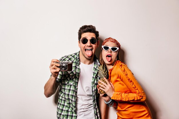 Young mischievous boy and girl show tongues on white background and hold pineapple and retro camera