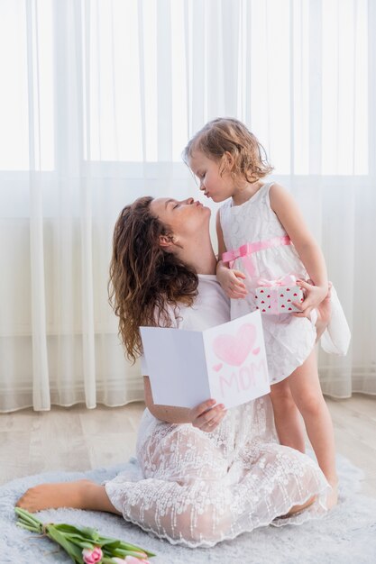 Young mother and daughter kissing at home with holding greeting card