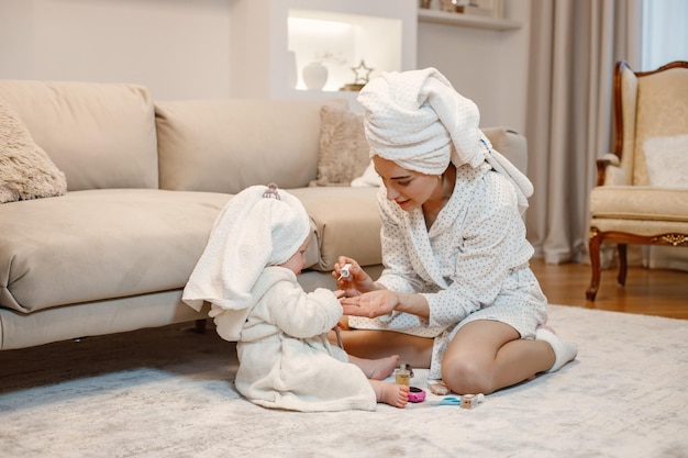 Free photo young mother and little baby girl wearing dressing gowns with hair wrapped in towels woman and girl sitting on a floor near sofa woman painting a nails of her daughter