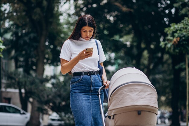 Young mother walking in park with baby stroller and talking on phone