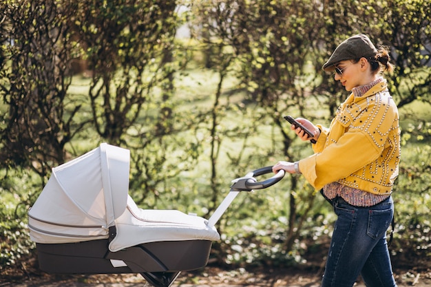 Young mother walking with baby carriage in park and using phone