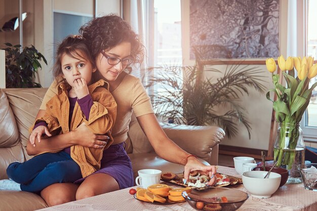 Young mother with cute little daughter having breakfast on the sofa at home.