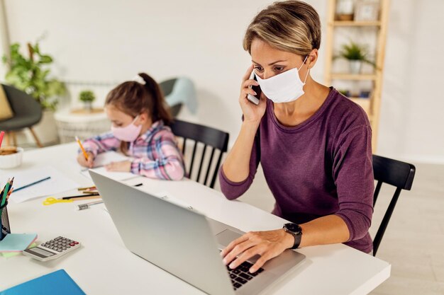 Young mother with protective face talking on the phone while working on laptop at home