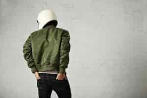 Free photo young motorcyclist in white helmet and green jacket portrait from the back with his hands in the back pockets of his jeans with white walls.