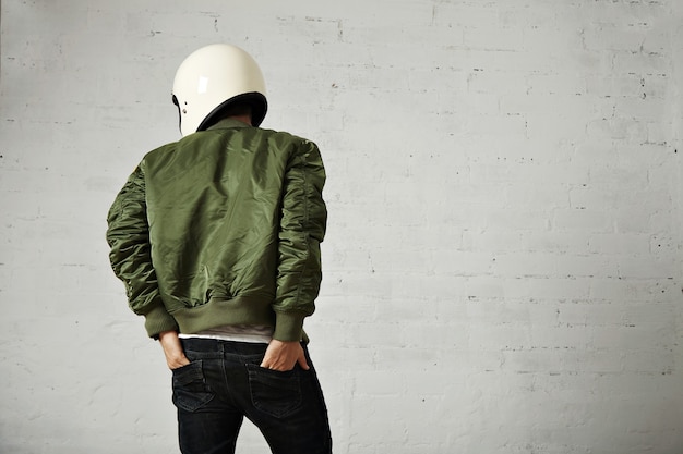 Free Photo young motorcyclist in white helmet and green jacket portrait from the back with his hands in the back pockets of his jeans with white walls.