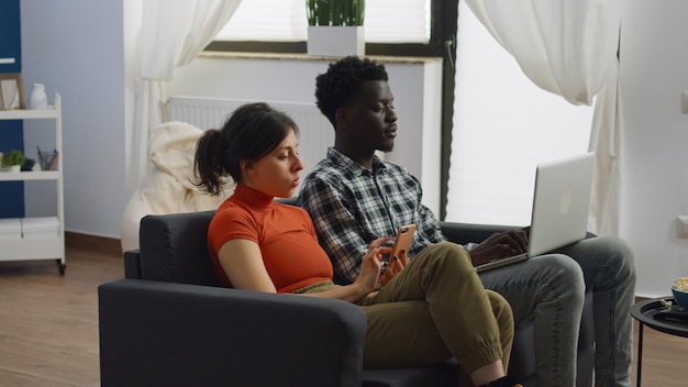 Young multi ethnic couple using technology in living room at home. Interracial people with smartphone and laptop while sitting together on couch. Mixed race wife and husband with devices