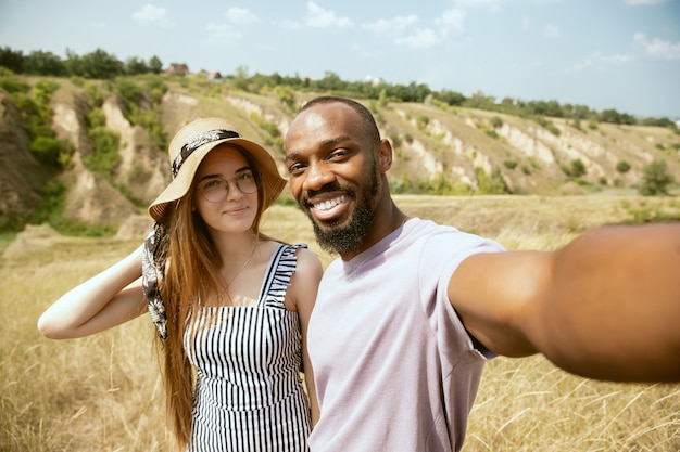 Young multiethnic international couple outdoors at the meadow in sunny summer day. African-american man and caucasian woman having picnic together. Concept of relationship, summertime. Making selfie.