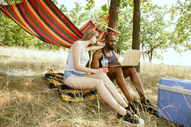 Young multiethnic international romantic couple outdoors at the meadow in sunny summer day. African-american man and caucasian woman having picnic together. Concept of relationship, summertime.