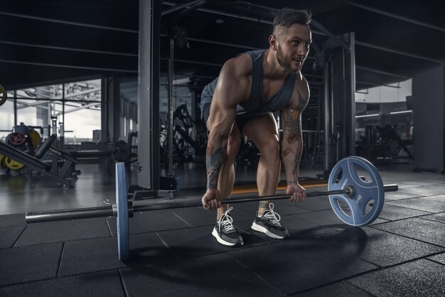 Young muscular athlete practicing pull-ups in gym with barbell