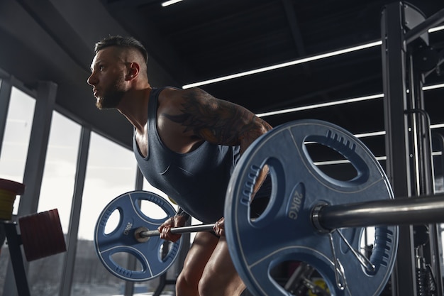 Young muscular athlete practicing pull-ups in gym with barbell