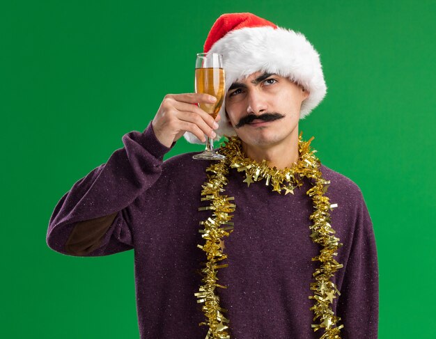 Young mustachioed man wearing christmas santa hat with tinsel around his neck holding glass of champagne  looking up puzzled 