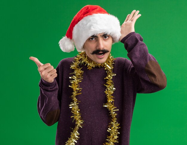 young mustachioed man wearing christmas santa hat with tinsel around his neck looking at camera surprised with arm raised pointing back with thumb standing over green background