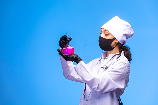 Young nurse in face and hand mask holds a test flask and follows the chemical reaction on blue wall