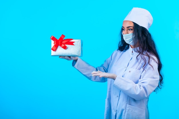 Young nurse in isolated uniform holding a gift box with red ribbon and presenting it.