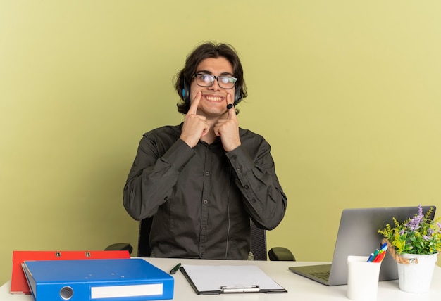 Free Photo young office worker man on headphones in optical glasses sits at desk with office tools using laptop holds smile with fingers isolated on green background with copy space