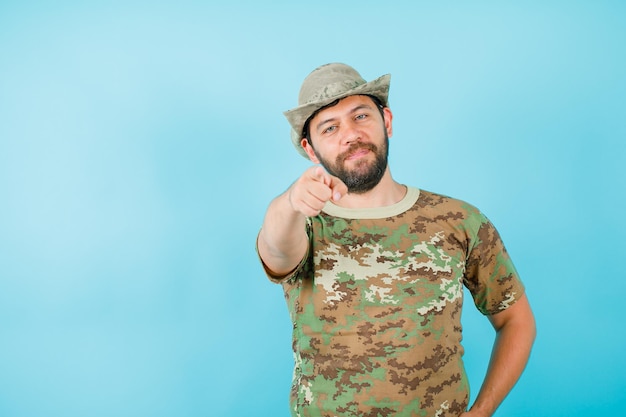 Free photo young officer is pointing camera with forefinger and putting other hand on waist on blue background