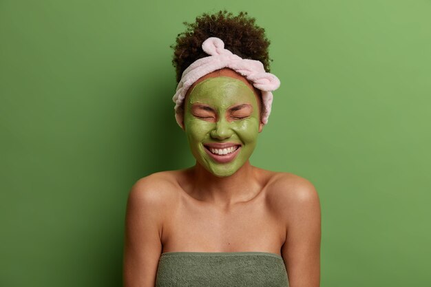 Young overjoyed woman applies natural clay mask for reducing acnes, smiles broadly, has perfect white teeth, has combed curly hair, headband, shows bare shoulders, isolated on green wall