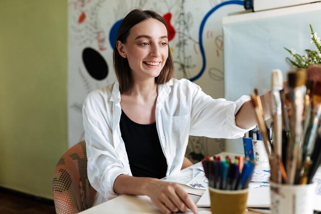 Young painter at the desk joyfully choosing paint brush with big pattern canvas on background at home