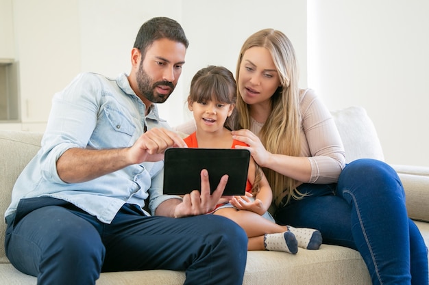 Free photo young parents couple and cute daughter sitting on couch, using tablet for video call or movie watching.