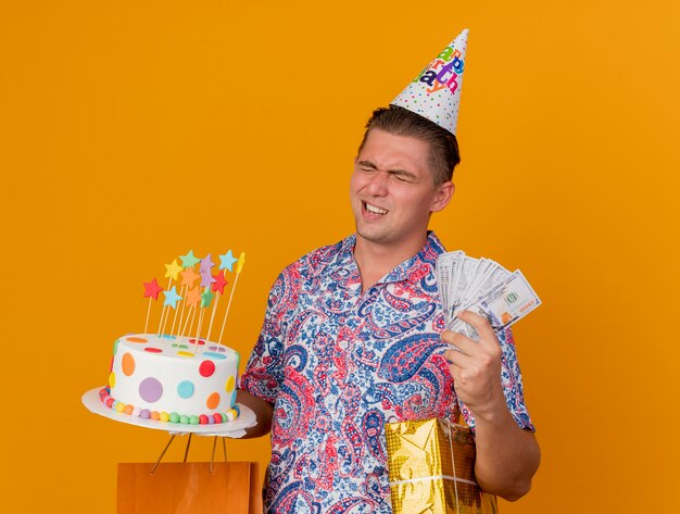 Young party guy wearing with closed eyes birthday cap holding gifts with cake and cash isolated on orange