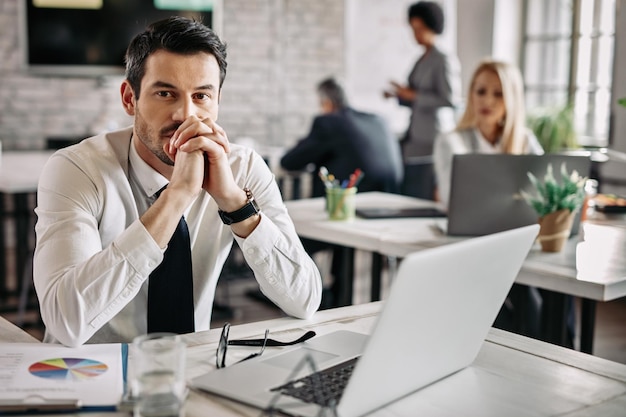 Young pensive businessman working at desk in the office and thinking of something There are people in the background