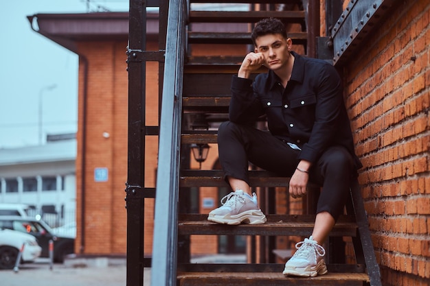 A young pensive guy wearing a shirt sitting on stairs outside near a building with the industrial exterior.