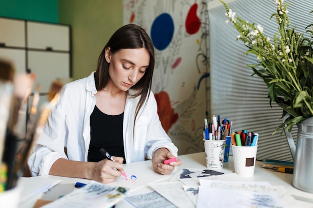 Free Photo young pensive painter at the desk dreamily drawing with pink marker while spending time in creative workplace at home