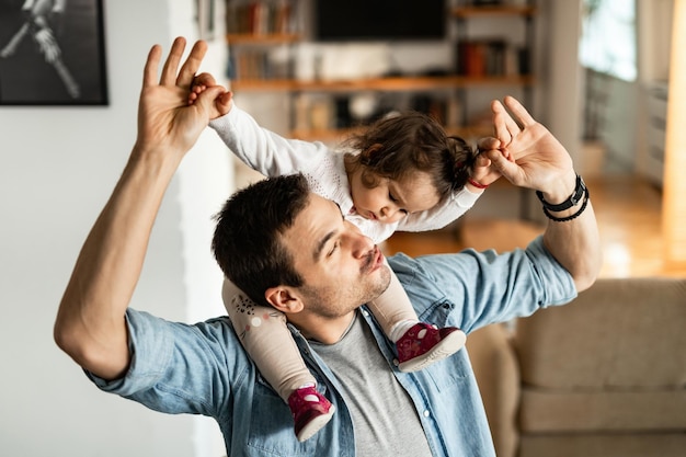 Young playful father having fun while carrying his small daughter on the shoulders at home