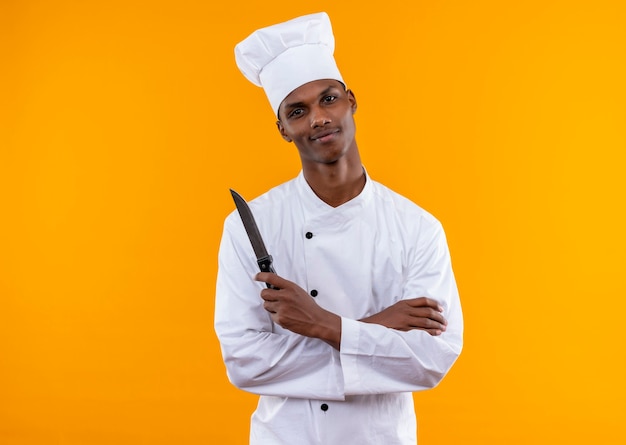 Young pleased afro-american cook in chef uniform crosses arms and holds knife isolated on orange wall