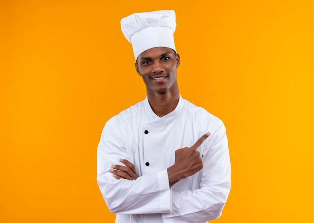 Young pleased afro-american cook in chef uniform crosses arms and points to the side isolated on orange wall