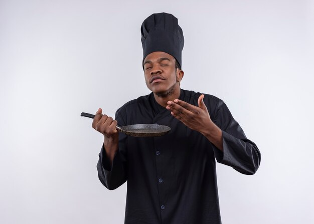 Young pleased afro-american cook in chef uniform holds frying pan and pretends to smell isolated on white wall 