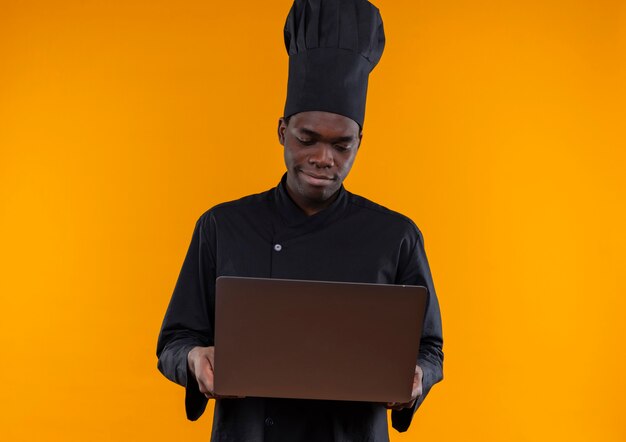 Young pleased afro-american cook in chef uniform holds and looks at notebook on orange  with copy space