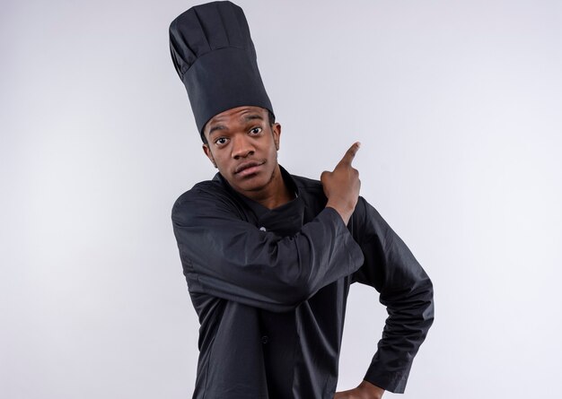 Young pleased afro-american cook in chef uniform points back isolated on white wall 