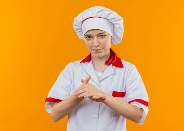 Young pleased blonde female chef in chef uniform holds hands together isolated on orange wall