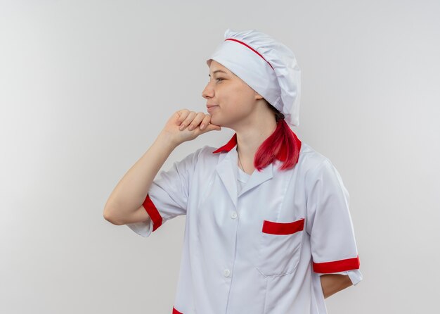 Young pleased blonde female chef in chef uniform looks at side and puts hand on chin isolated on white wall