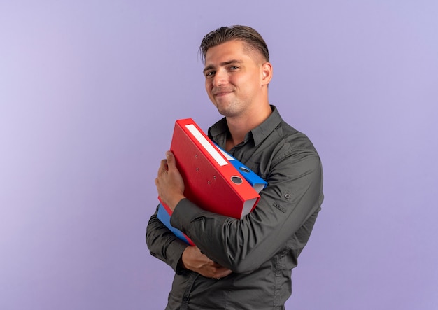 Free photo young pleased blonde handsome man holds file folders isolated on violet space with copy space