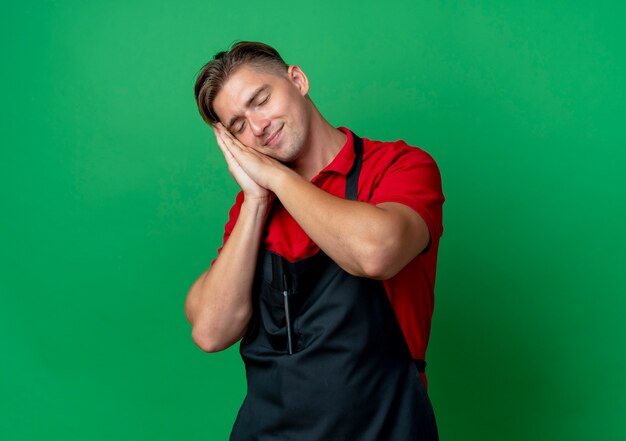 Young pleased blonde male barber in uniform pretends to sleep putting head on hands isolated on green space with copy space