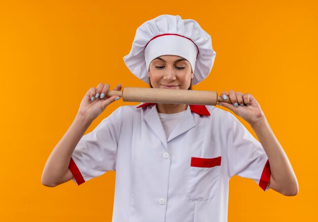 Young pleased caucasian cook girl in chef uniform holds and looks at rolling pin isolated on orange space with copy space