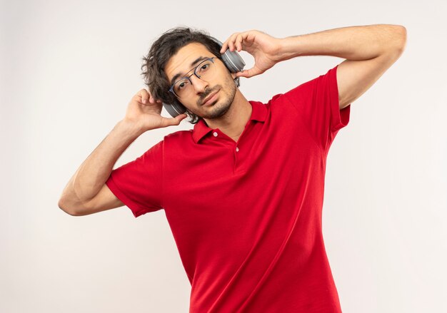 Young pleased man in red shirt with optical glasses puts on and holds headphones isolated on white wall