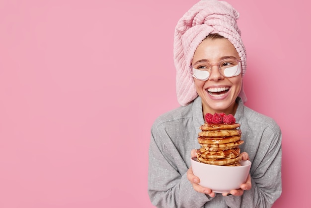 Free Photo young positive woman holds bowl with pile of delicious pancakes smiles toothily concentrated away applies beauty patches to reduce wrinkles feels refreshed after sleeping wears pajama stands indoor