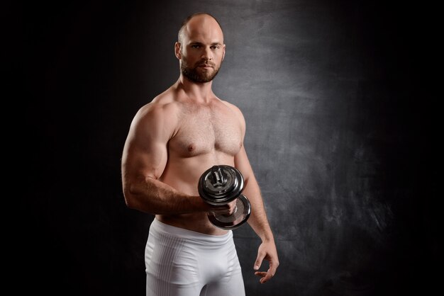 Young powerful sportsman training with dumbbells over black wall.