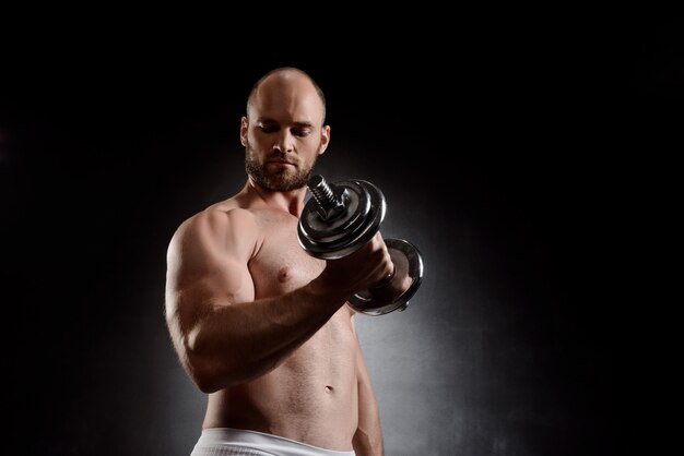 Young powerful sportsman training with dumbbells over black wall.