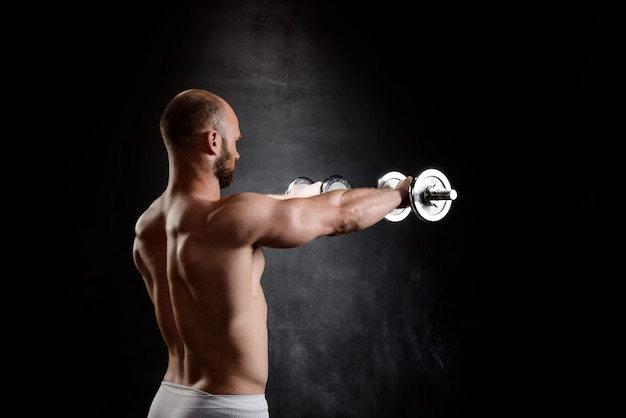 Young powerful sportsman training with dumbbells over black wall.