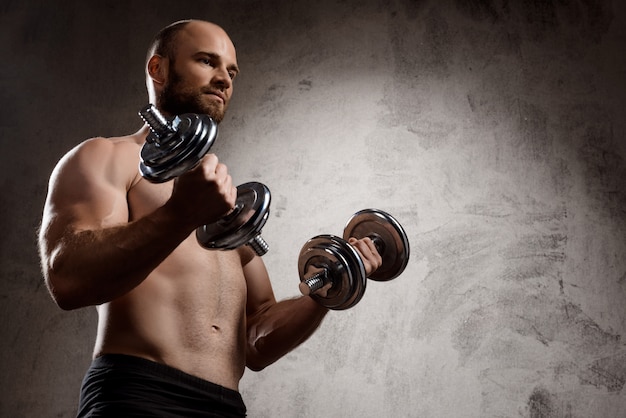 Young powerful sportsman training with dumbbells over dark wall.