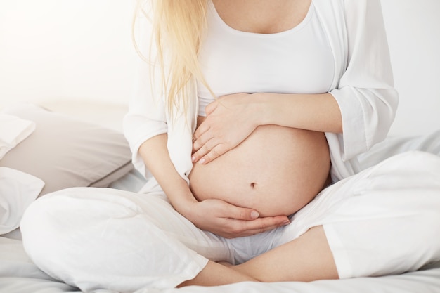 Young pregnant soon to be mother touching her belly sitting in her bedroom in afternoon. Pregnancy concept.