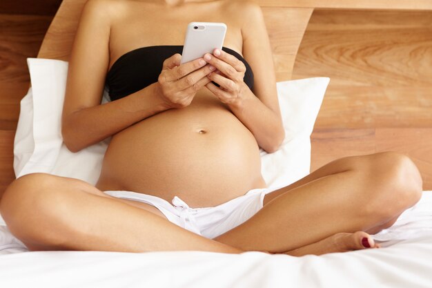 Young pregnant woman in underwear relaxing on her bed in the morning viewing pictures online on Internet using electronic gadget. Cropped view of female expecting baby reading e-book on smart phone