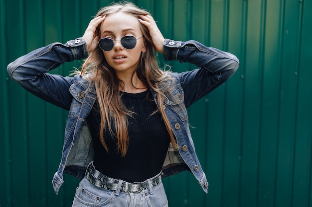 Young pretty attractive girl in denim clothes in glasses on a simple dark green wall with place for text.